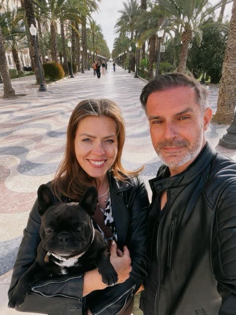Marek with his wife and their French bulldog on a palm-lined promenade in Spain